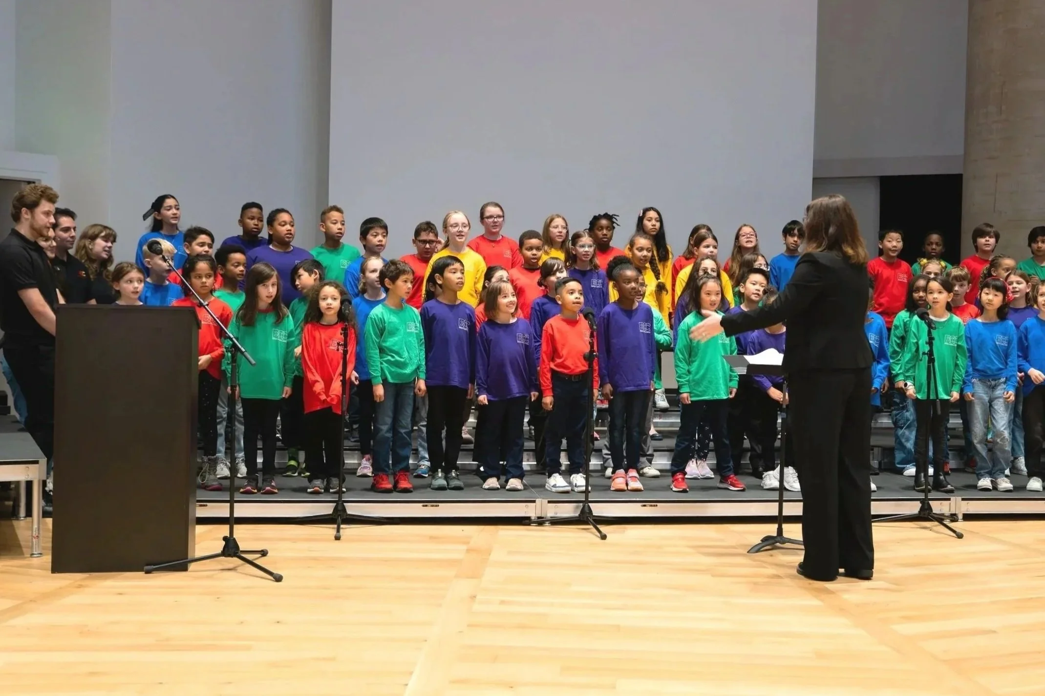 A choir of children wearing brightly colored shirts stands on risers on a stage, directed by a woman in a black blazer, with a man and a woman playing piano on the side.