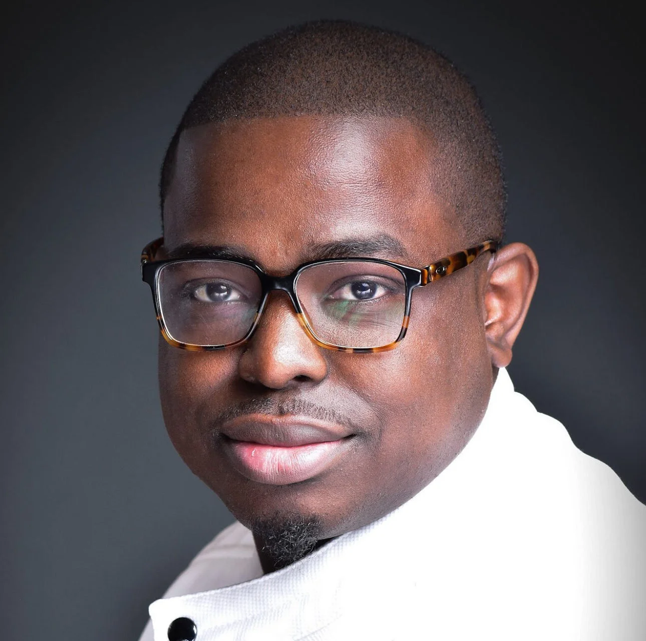 Close-up portrait of a young Black man wearing tortoiseshell glasses and a white shirt, smiling slightly against a dark background.