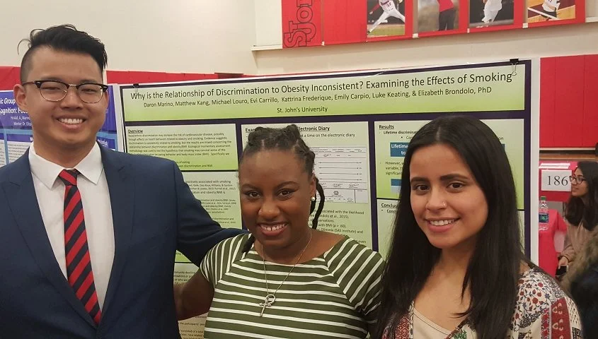 Three students standing in front of a science research poster at a conference, smiling at the camera. Two women and one man, with the man wearing glasses and a suit with a red and black striped tie.