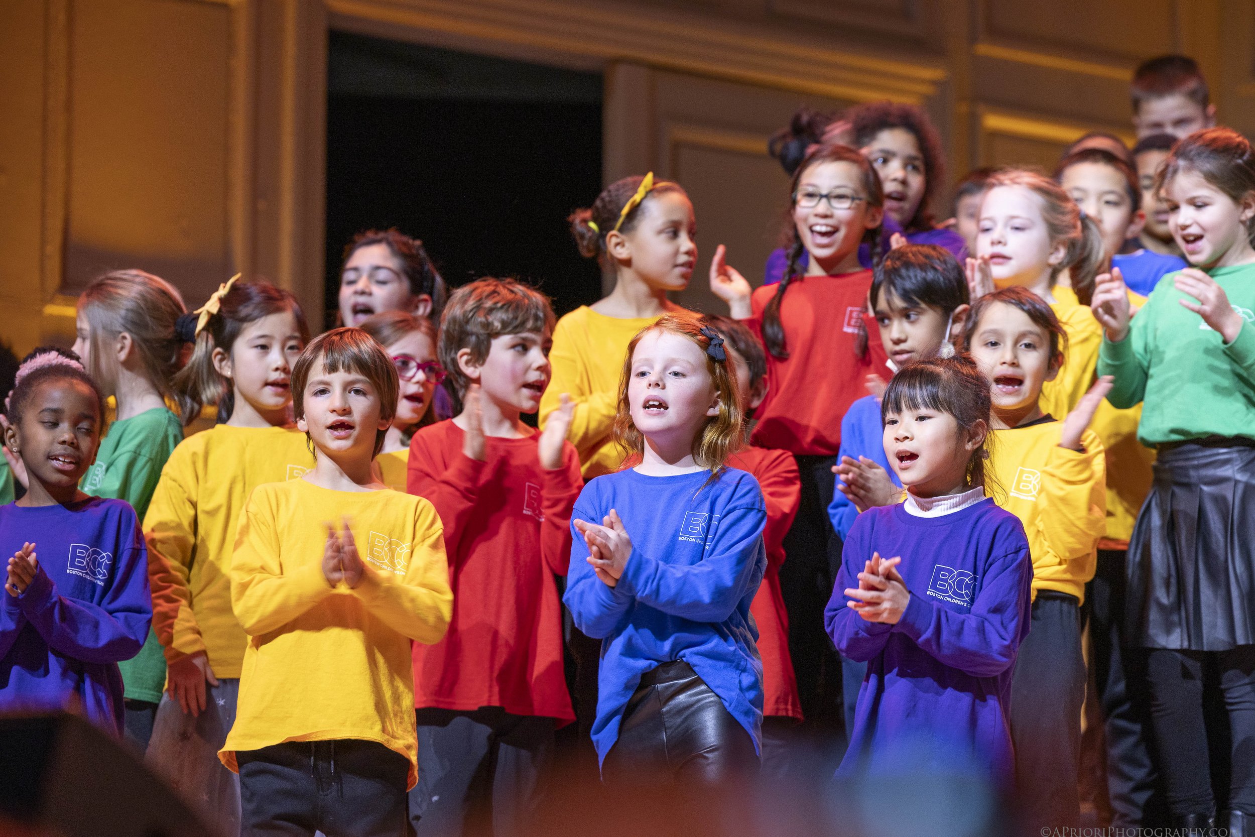 A group of children on stage, dressed in colorful shirts, singing or performing together at a children's event.