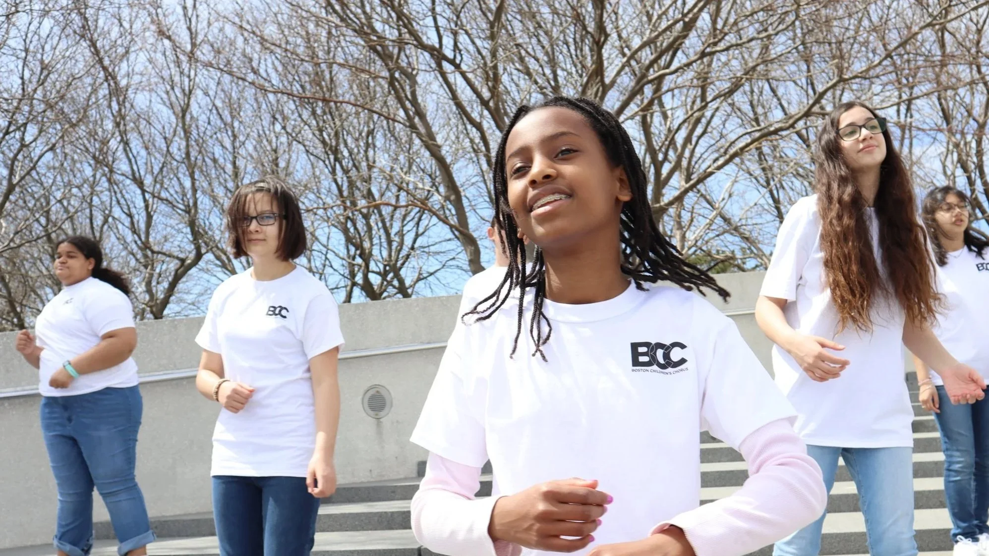 Group of children wearing white t-shirts with Boston Children's Chorus logo, walking outside near stairs with leafless trees in the background.