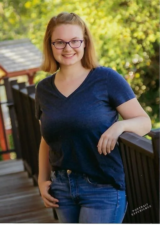 A young woman with glasses and shoulder-length hair smiling outdoors, leaning on a wooden railing with trees in the background.