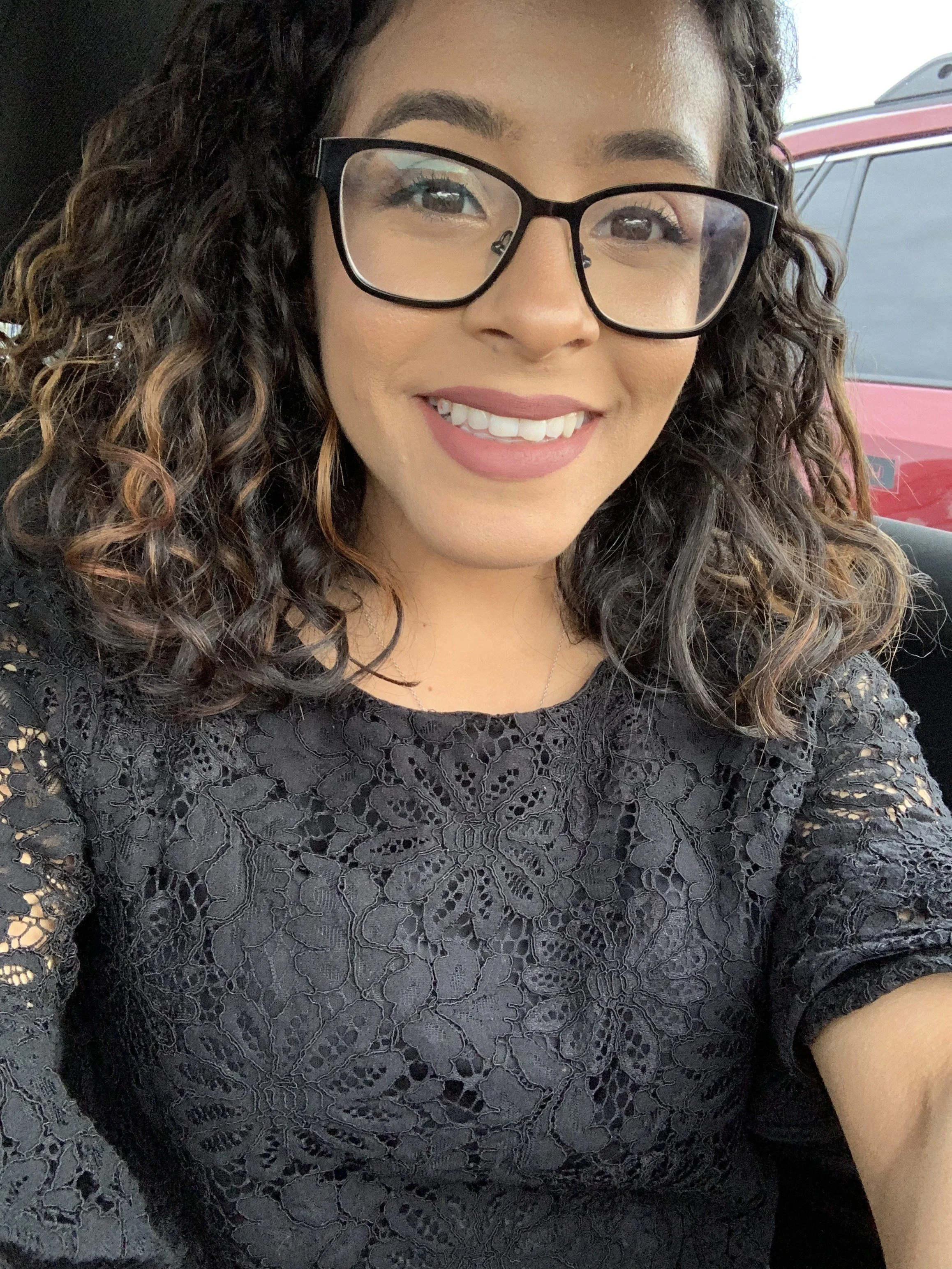 Close-up of a smiling woman with curly hair wearing glasses and a black lace top, sitting inside a vehicle with a red car visible outside the window.