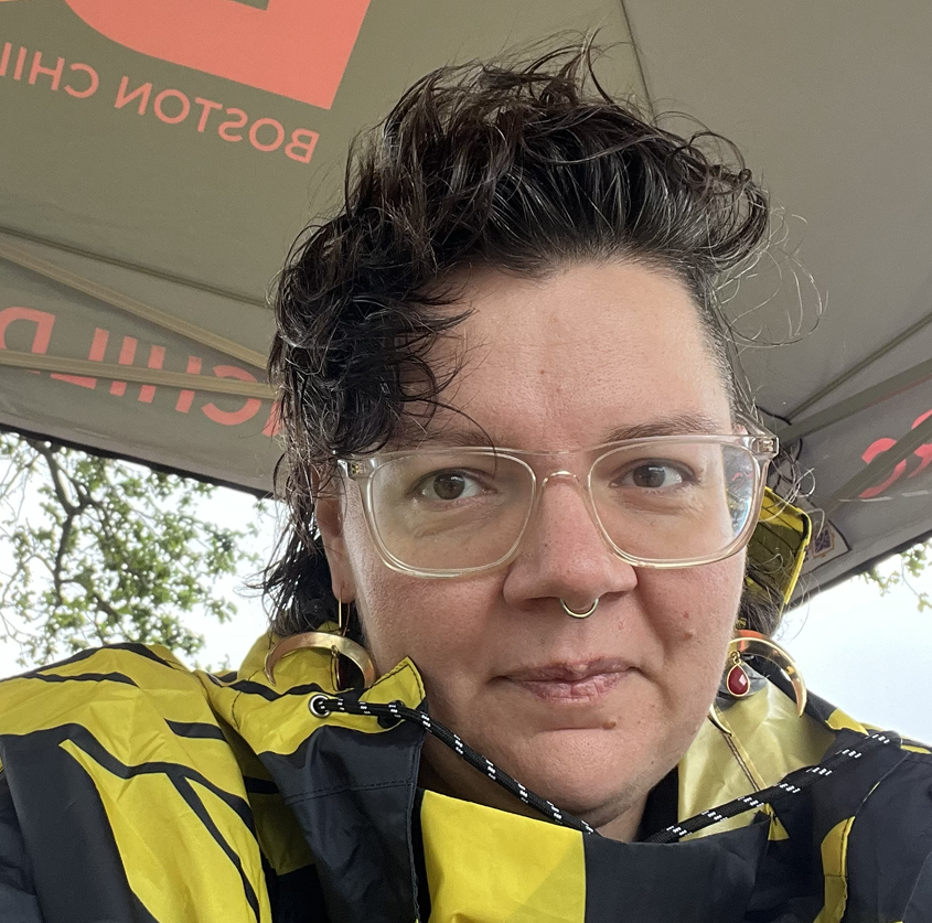 A person with short, curly dark hair, wearing clear glasses, a yellow and black rain jacket, and gold jewelry, sitting under a tent with the words "Boston Children" partially visible in the background.