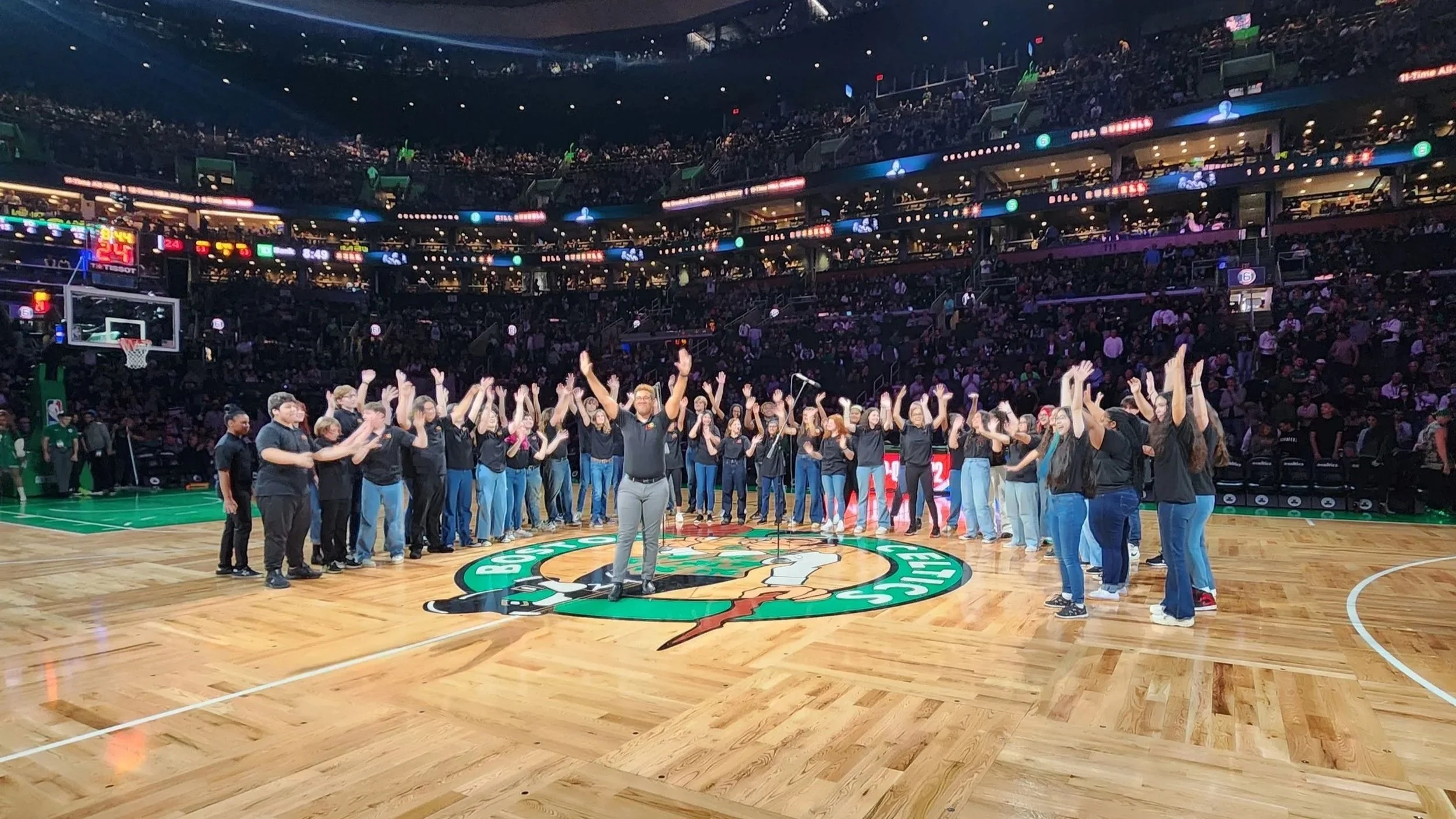 A group of people standing on a basketball court, clapping and raising their hands, with many spectators in the stands and a large Boston Celtics logo at center court.