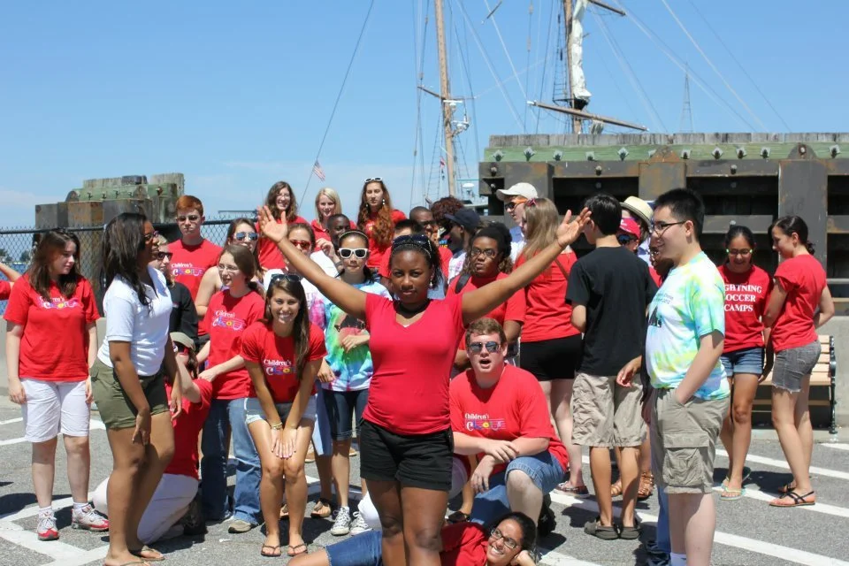 A diverse group of children and teenagers gathered outdoors on a sunny day, some wearing red shirts, with a woman in front with arms raised, and a ship in the background.