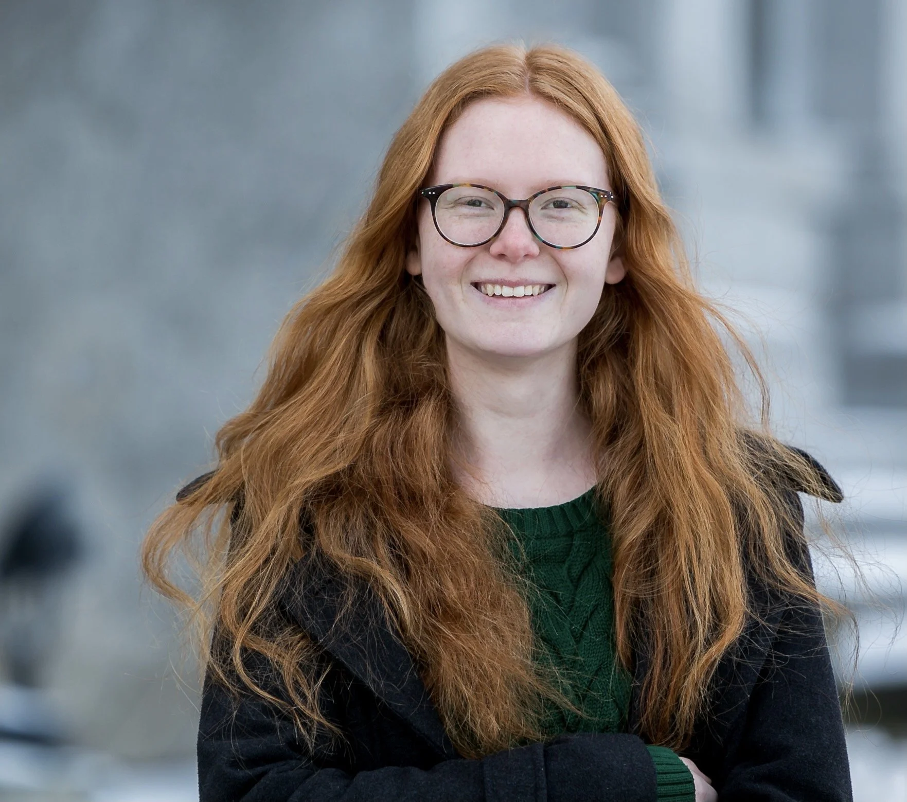 A woman with long, curly red hair, wearing glasses, smiling, dressed in a dark blazer and green sweater, standing outdoors with a blurred city background.