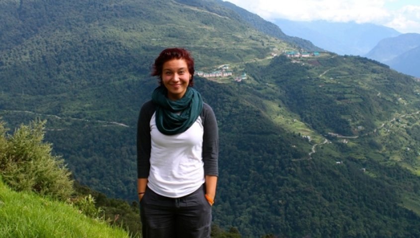 A woman with short dark hair, wearing a white and black long-sleeve shirt, standing outdoors with green hills and a mountain landscape in the background.