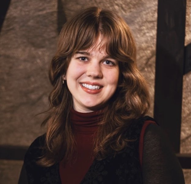 A young woman with shoulder-length wavy brown hair, wearing a maroon turtleneck and a black vest, smiling in front of a brick wall background.