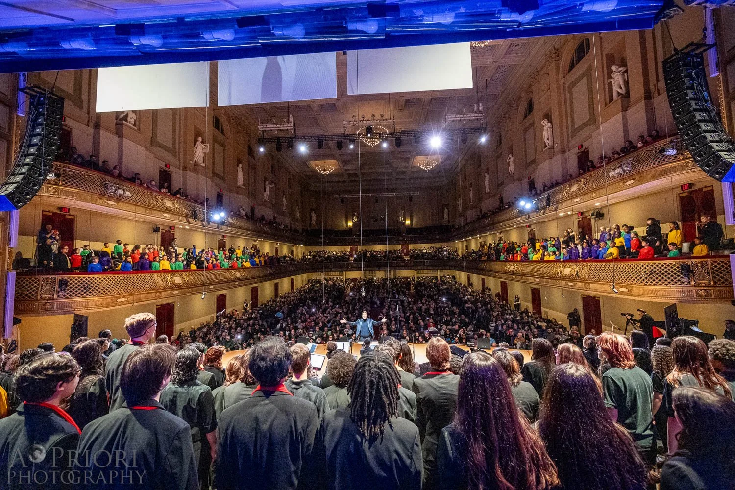 A large choir standing on stage, a conductor in front, in a grand theater with multiple balconies and ornate decor, filled with a large audience.