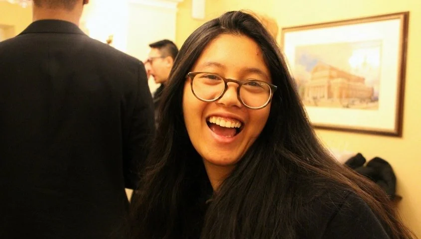 A young woman with long black hair and glasses smiling and laughing indoors, with framed artwork on the wall behind her.