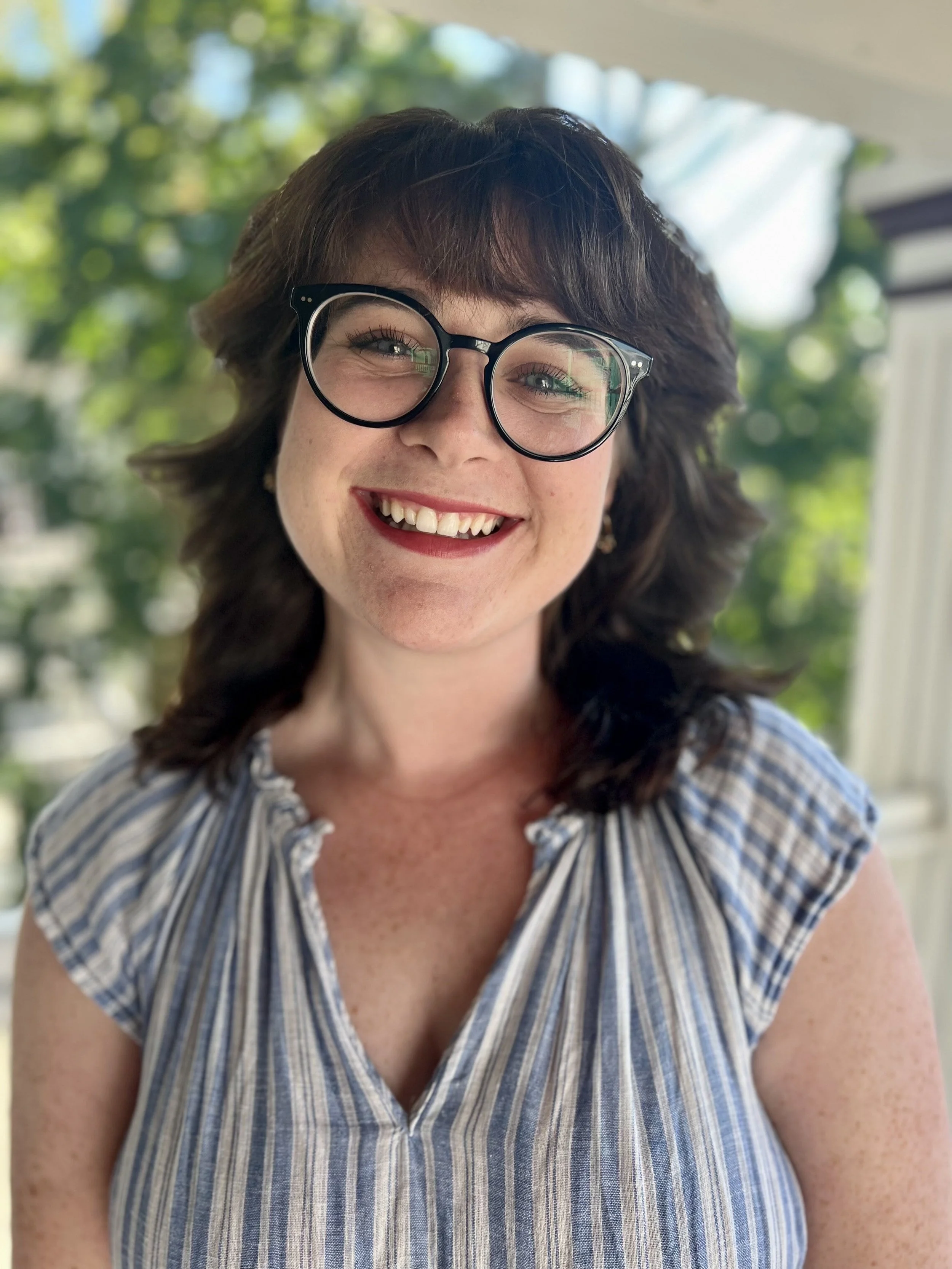 A woman with short brown hair and glasses smiling in front of a window with green trees outside.