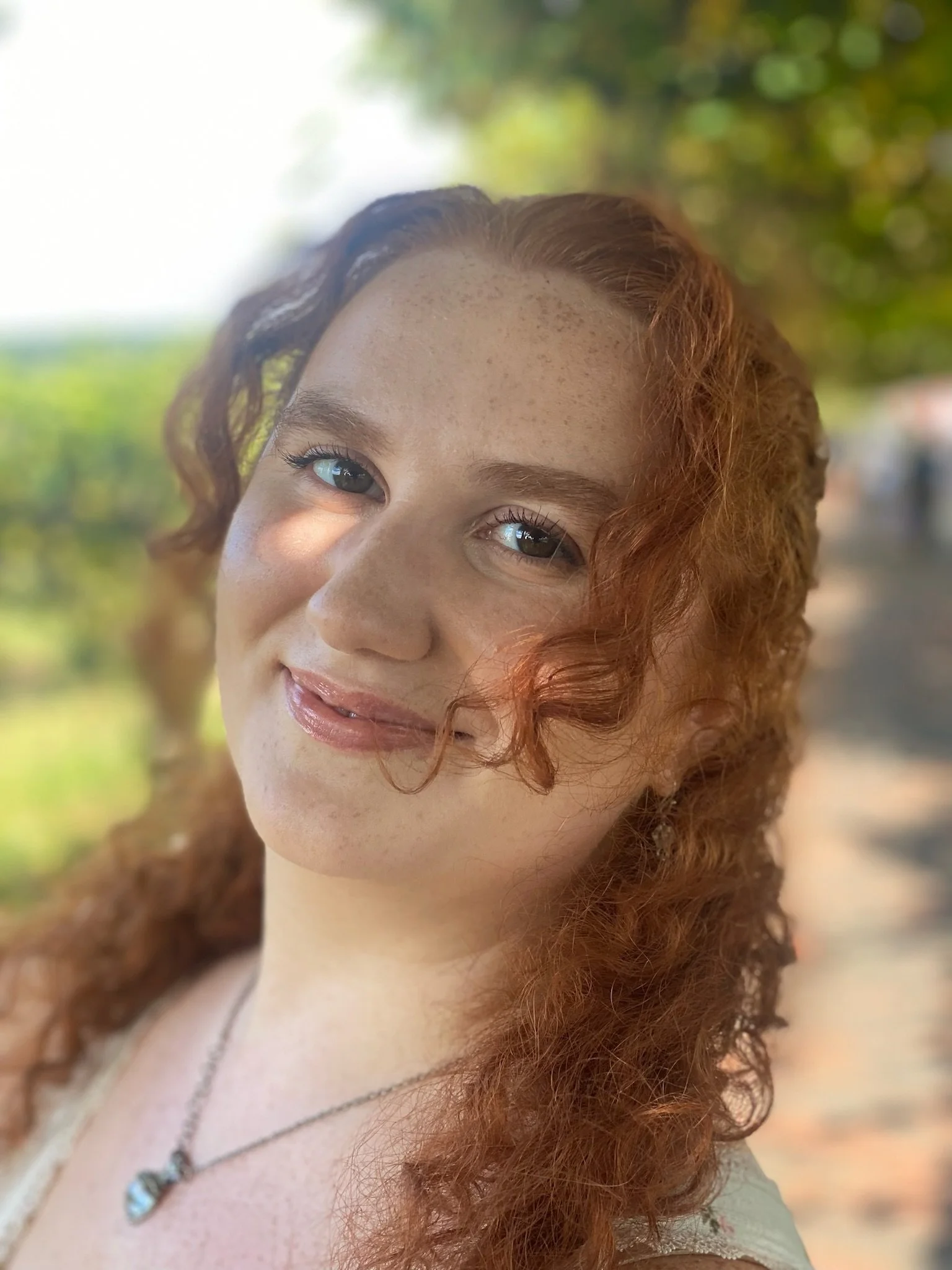 Close-up of a smiling woman with red curly hair, blue eyes, freckles, wearing necklaces, outdoors with blurred green foliage in the background.