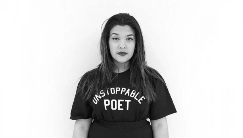 A Black and white photo of a young woman with shoulder-length hair, wearing a dark t-shirt that says 'UNSTOPPABLE POET.' She has a serious expression and is standing against a plain light background.