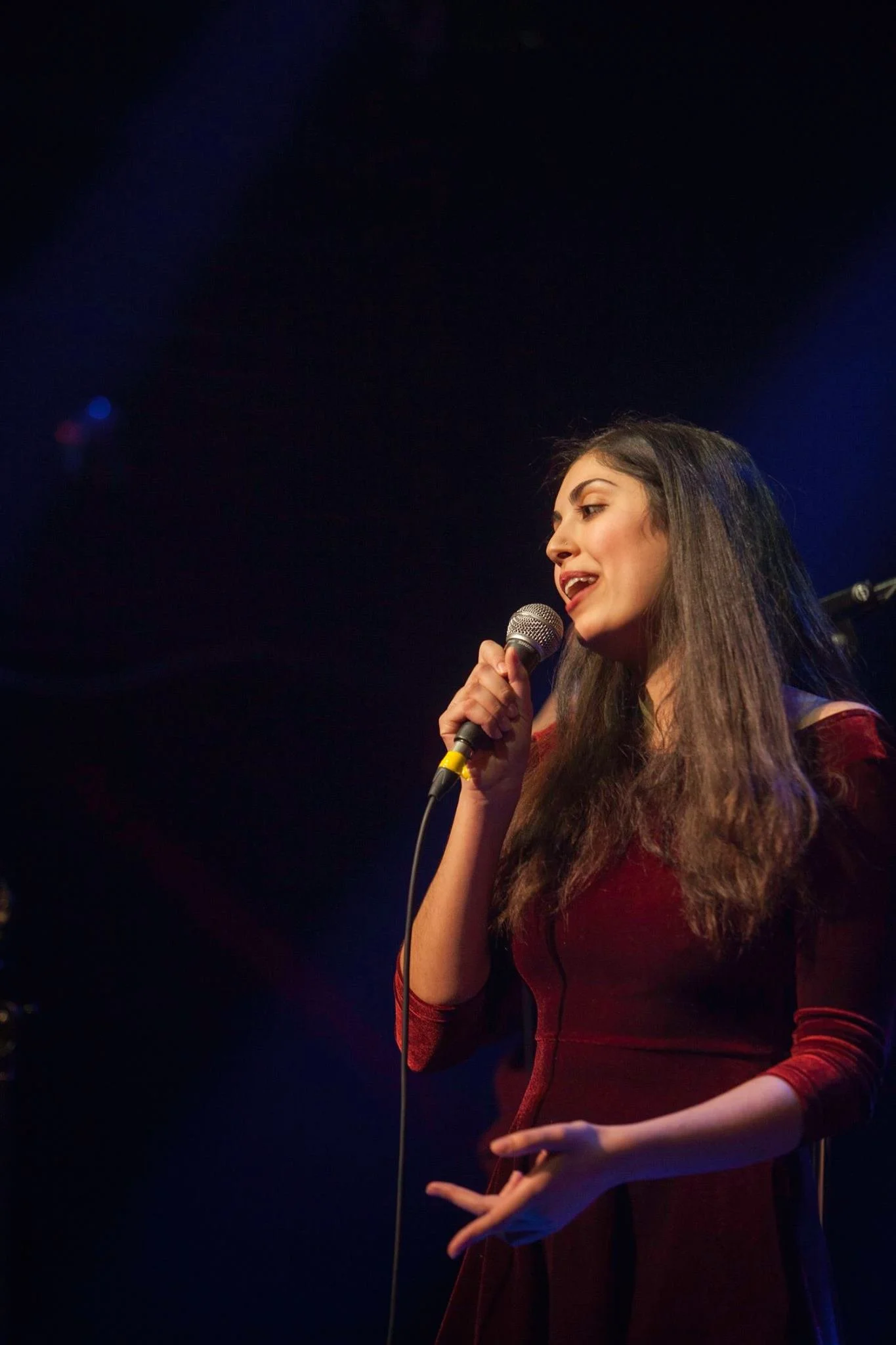 A young woman with long dark hair singing into a microphone on stage, wearing a red dress, with a dark background and stage lights.