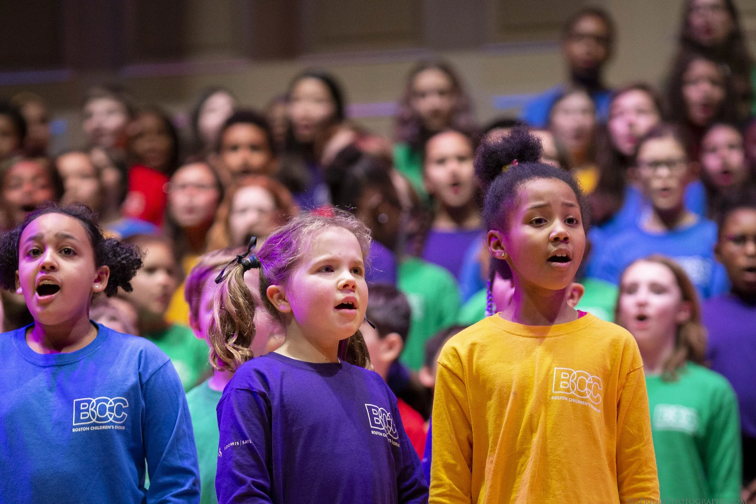 Young children, wearing colorful shirts with the Boston Children's Chorus logo, sing passionately in a choir.