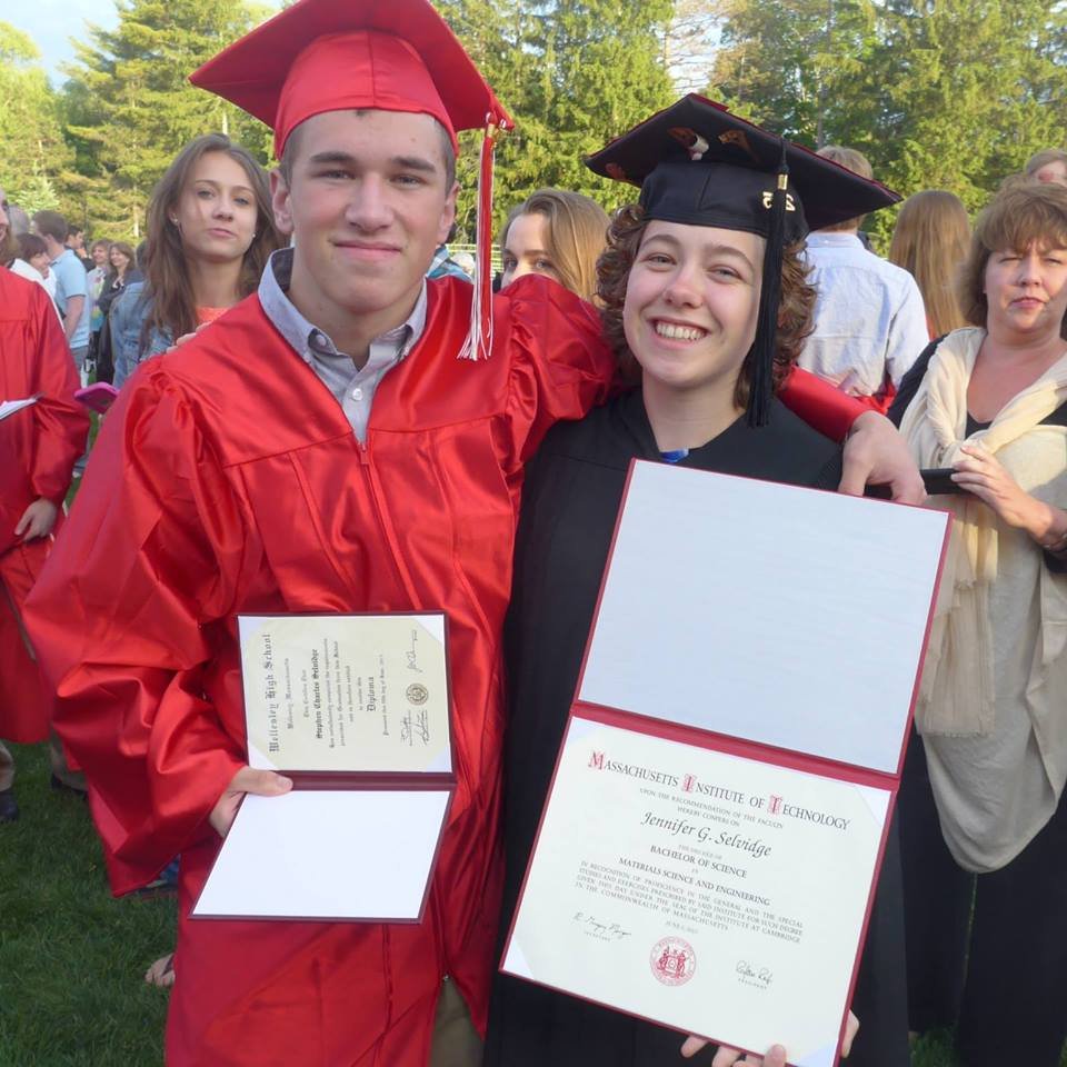 Two students in graduation caps and gowns, one in red and the other in black, smiling and holding diplomas at an outdoor graduation ceremony.