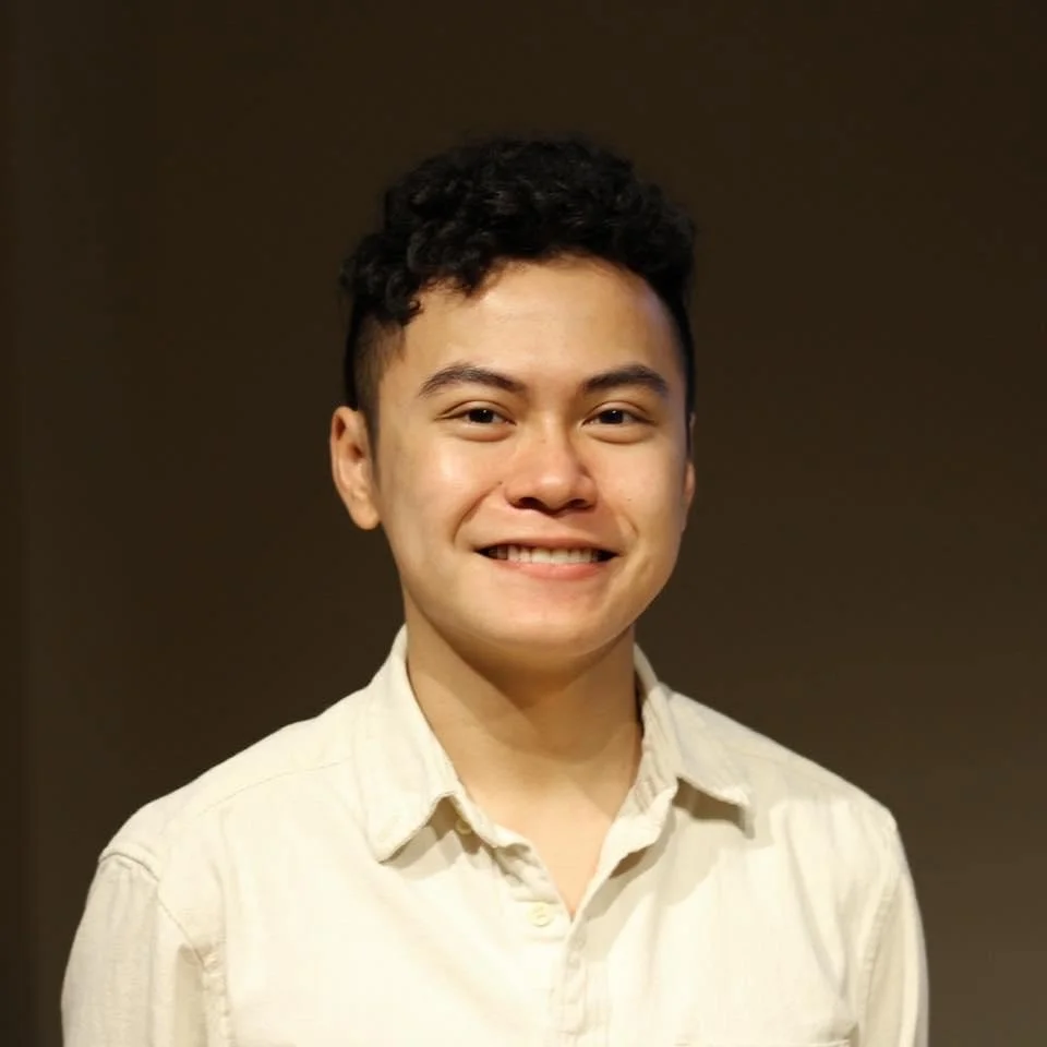 A young man with dark curly hair and a light beige button-up shirt smiling at the camera against a plain dark background.