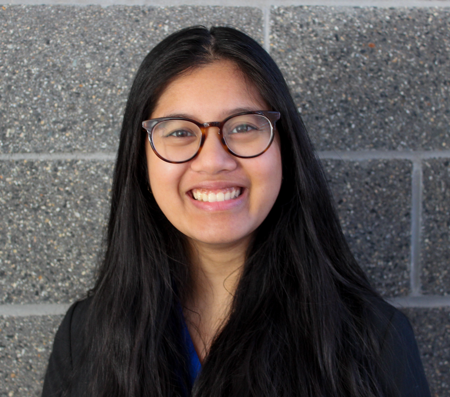 A woman with long black hair, glasses, and a bright smile, wearing a black jacket and standing against a gray brick wall.