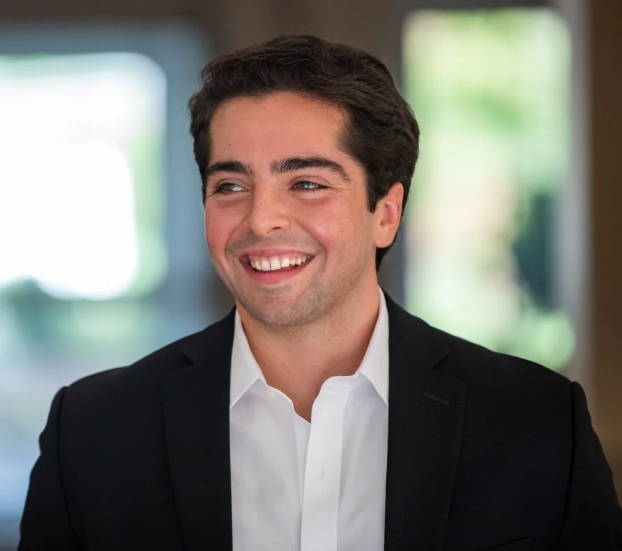 A young man with dark hair and a bright smile, dressed in a black suit and white shirt, standing indoors with blurred windows and greenery in the background.
