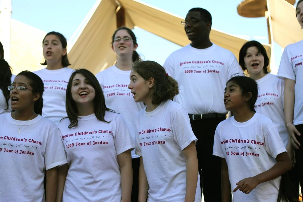 Children and adults singing together outdoors, wearing matching white T-shirts with red text, during a choir event titled '2009 Tour of Jordan'.