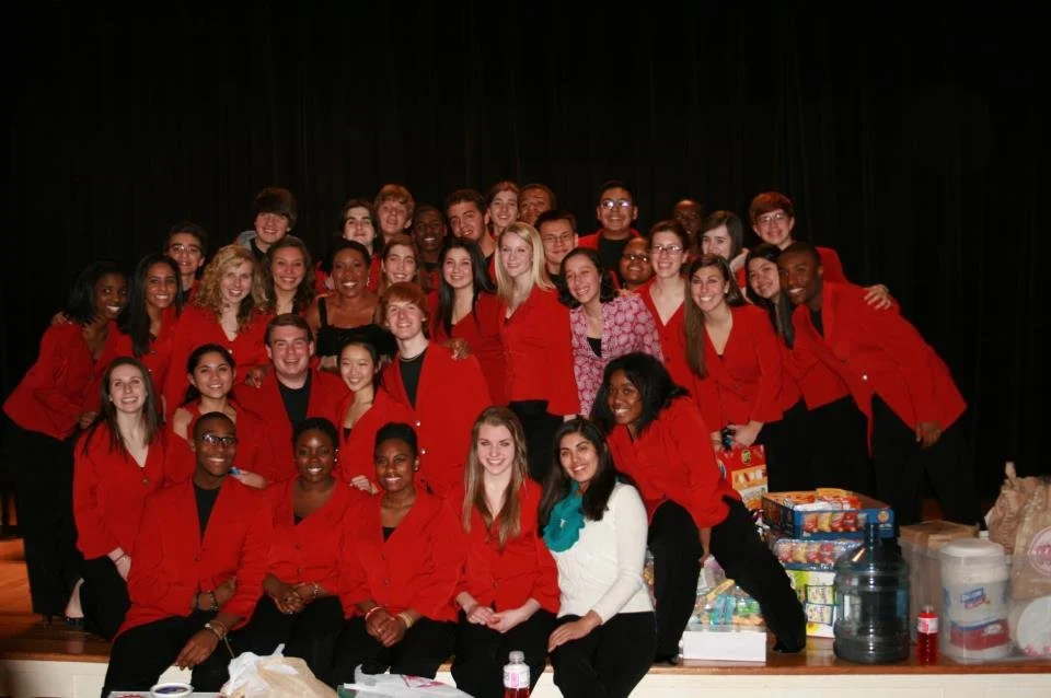 Group of young adults, many wearing red blazers, gathered on a stage with black curtains background, celebrating with boxes of food and drinks.
