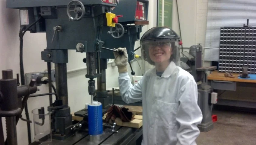 A woman in a white lab coat, safety goggles, and a black helmet in a workshop. She is smiling and holding a large drill press handle, with various tools and equipment around her.