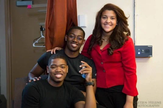 Three young adults smiling and posing together indoors, with one person sitting and two standing behind, in casual and business casual attire.