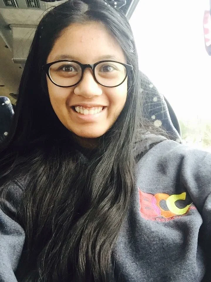 A smiling young woman with long dark hair, glasses, and a dark hoodie taking a selfie in a bus.