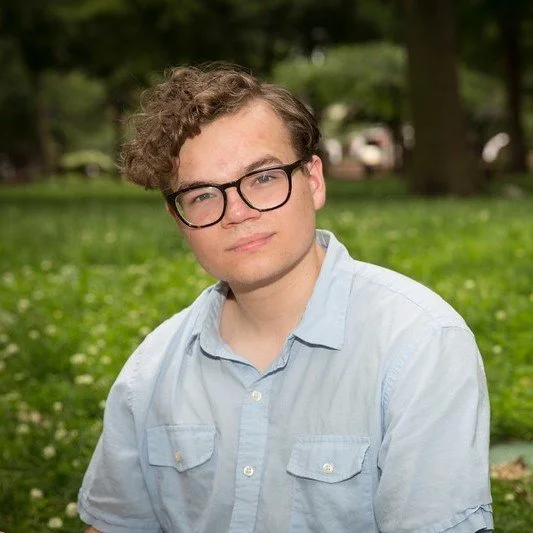 A young man with curly brown hair and glasses wearing a light blue shirt outdoors in a park with green grass and trees in the background.