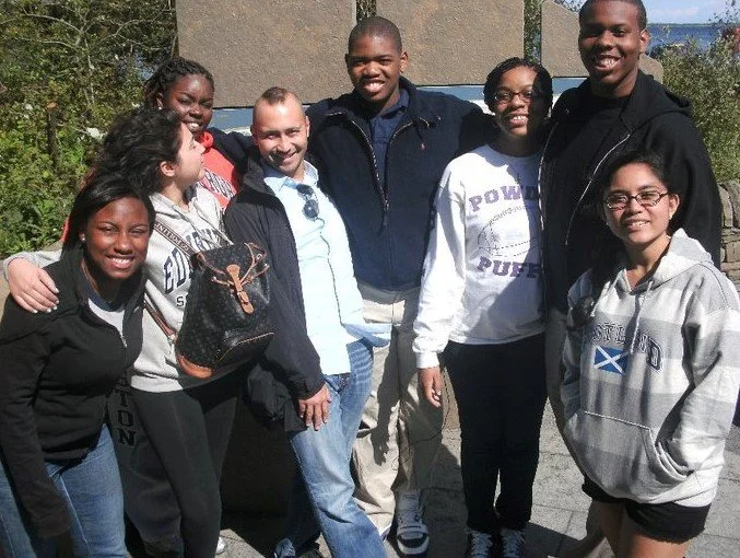 Group of nine young adults standing outdoors, smiling at the camera, with a mix of casual clothing and jackets.