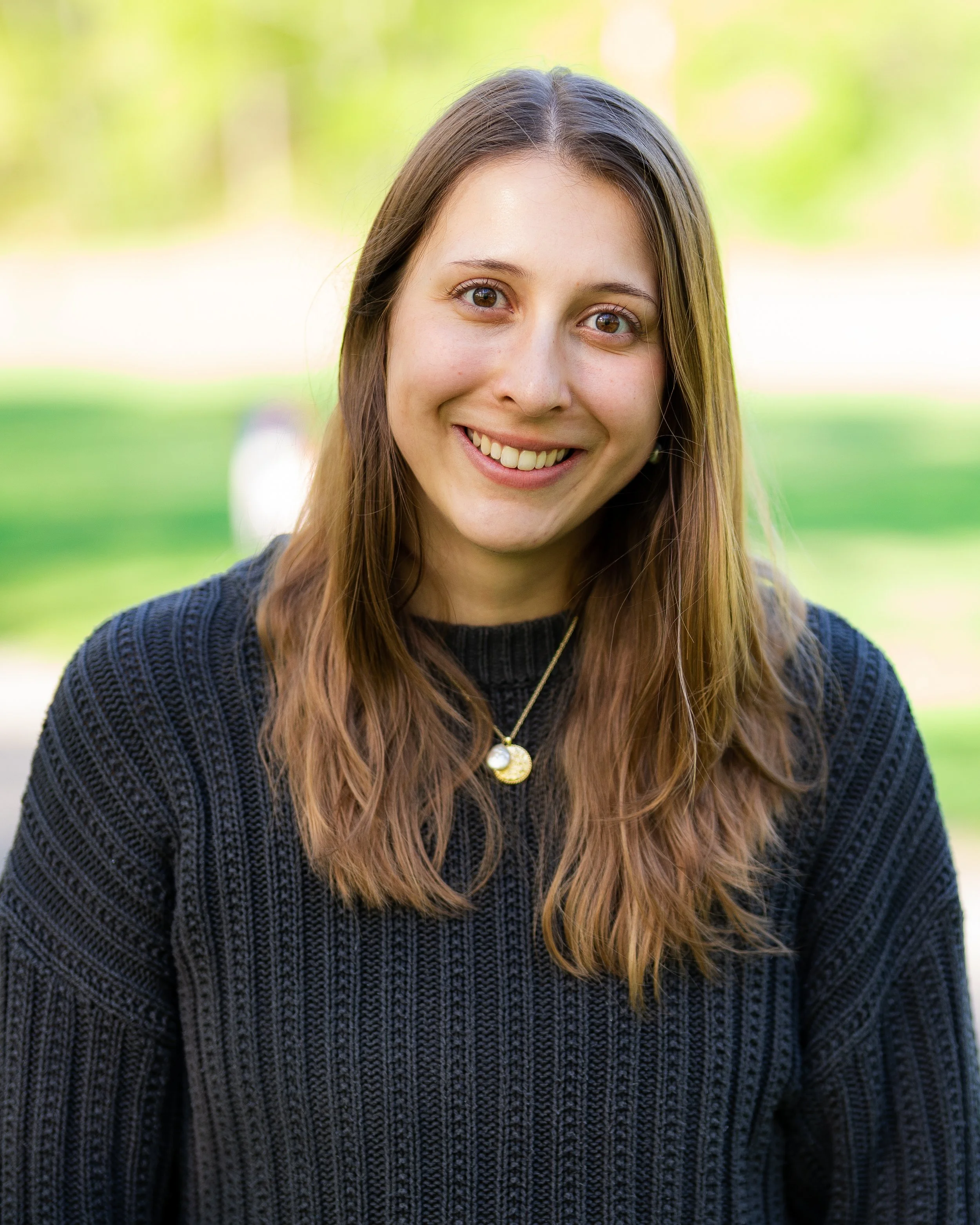 Portrait of a young woman with long brown hair, smiling outdoors. She is wearing a black sweater and a gold necklace with two pendants, one circular and one oval. The background is blurred with green foliage.