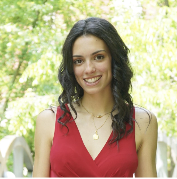 A smiling woman with dark wavy hair, wearing a red sleeveless dress and gold necklaces, outdoors with green trees in the background.
