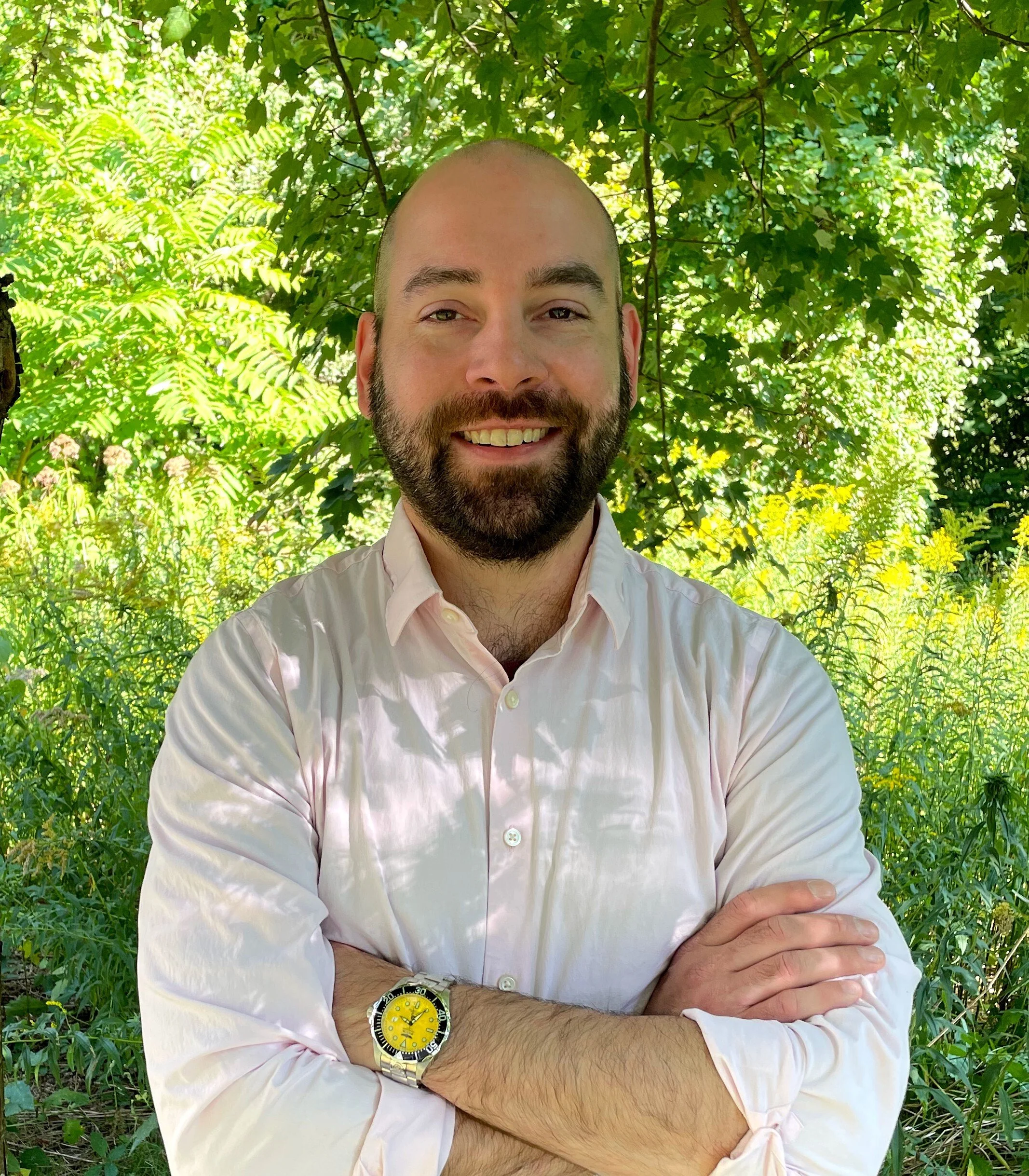 A man with a beard and bald head smiling, wearing a white shirt and a yellow watch, standing outdoors in front of lush green trees and plants with sunlight filtering through.