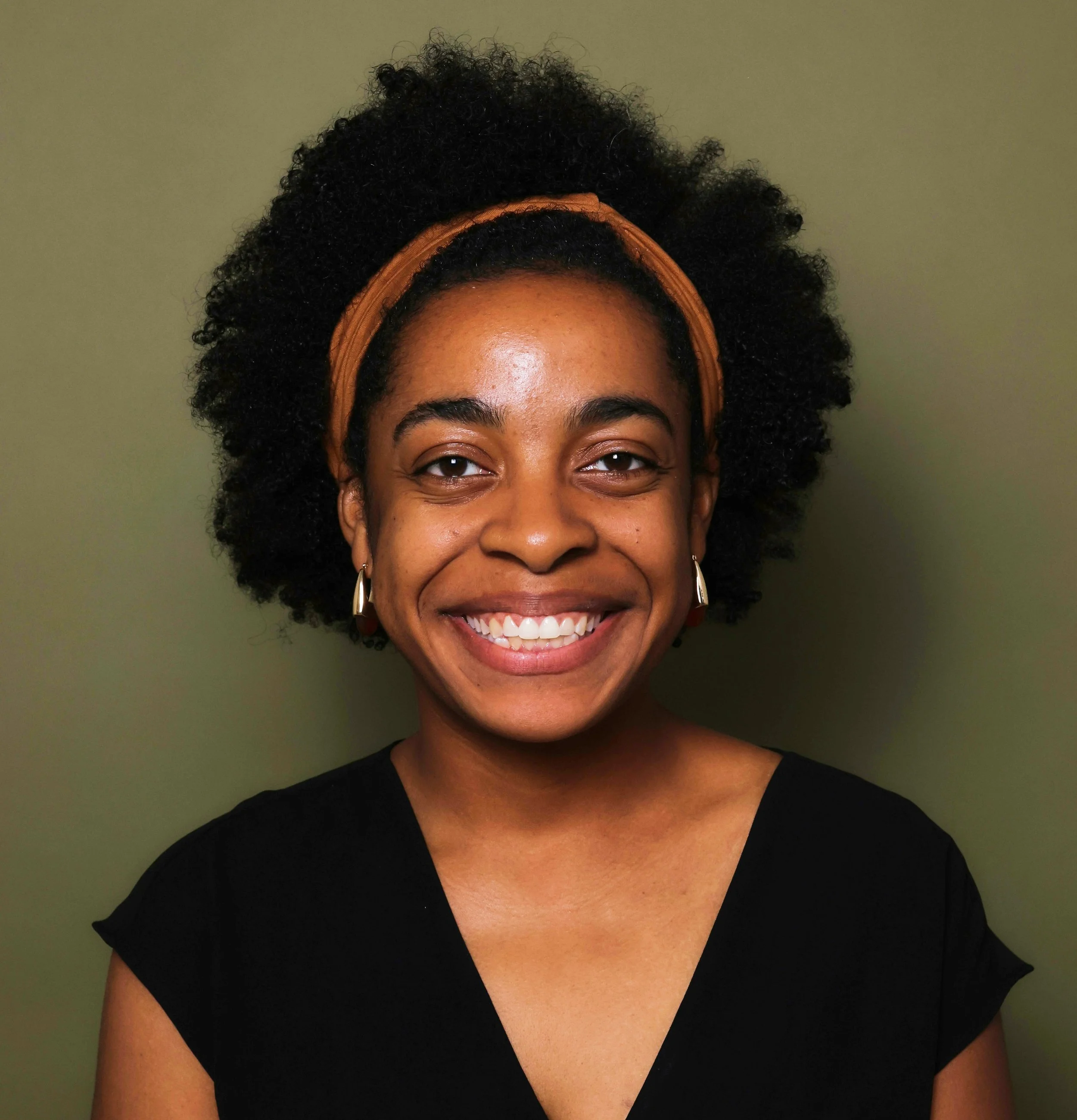 Close-up of a woman smiling with dark curly hair and wearing a brown headband and earrings, against a green background.