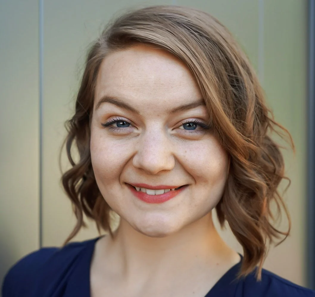 Close-up of a smiling young woman with wavy light brown hair and blue eyes, wearing a dark navy top, standing against a blurred light-colored background.