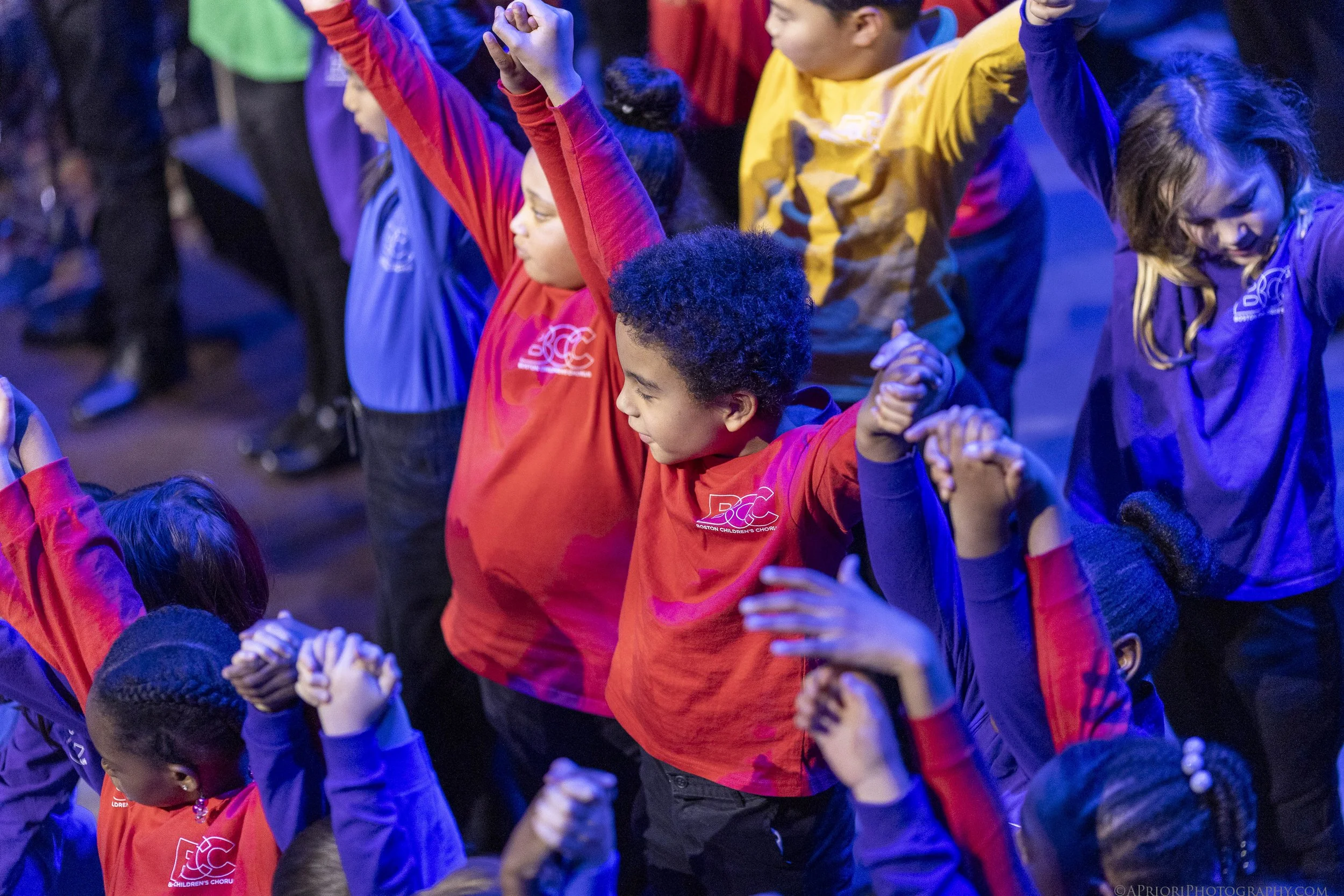 A group of children wearing colorful clothing, holding hands, and raising their arms in a close circle, possibly during a dance or group activity.