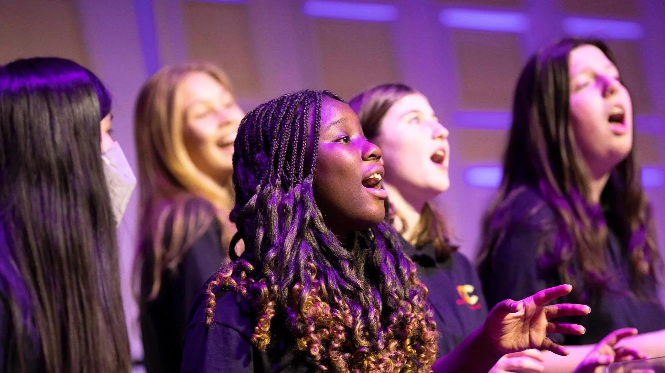 A diverse group of women singing in a choir, illuminated by purple stage lights.