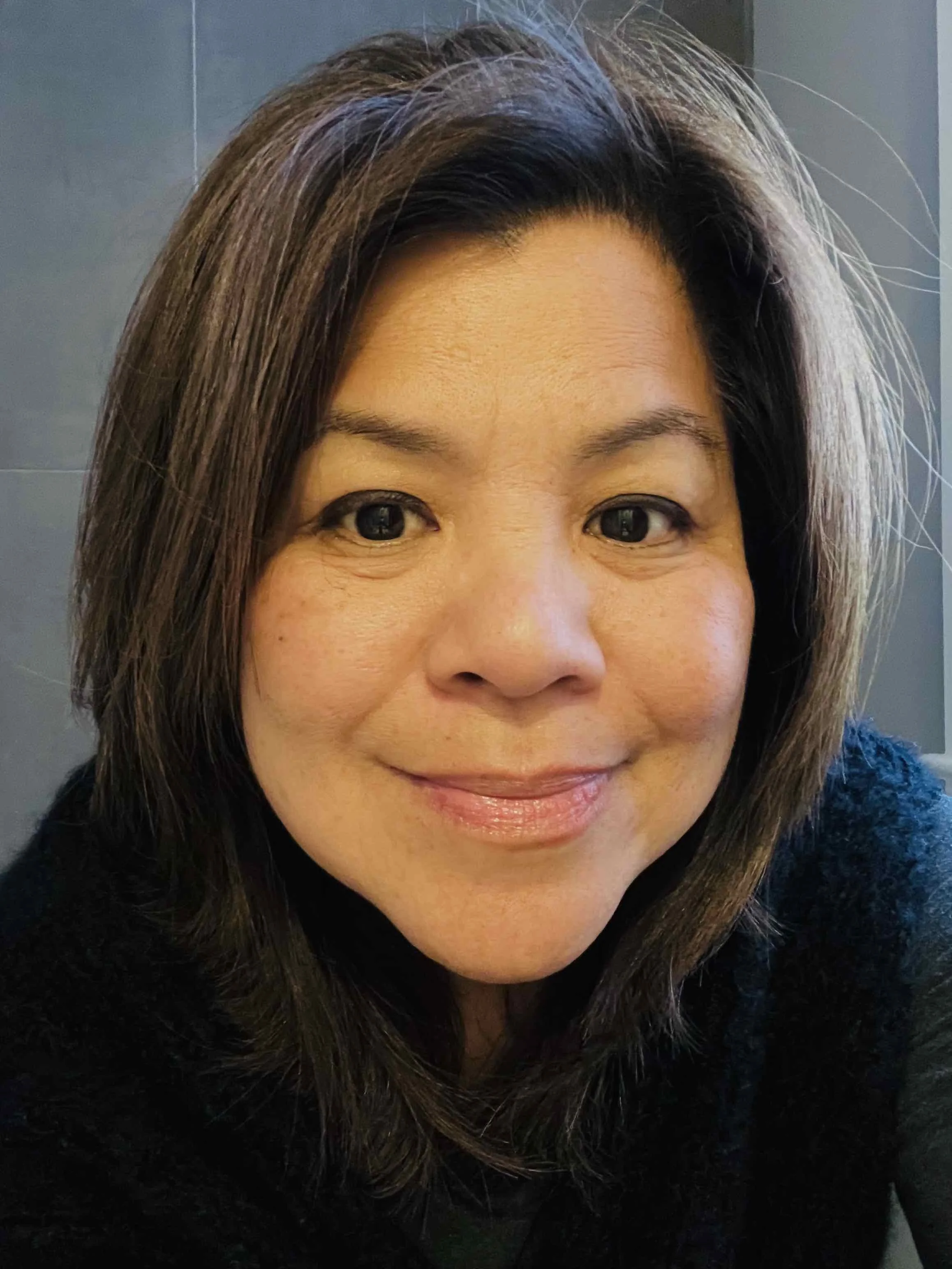 Close-up of an Asian woman with shoulder-length dark hair, smiling slightly, wearing a black top, against a plain background.