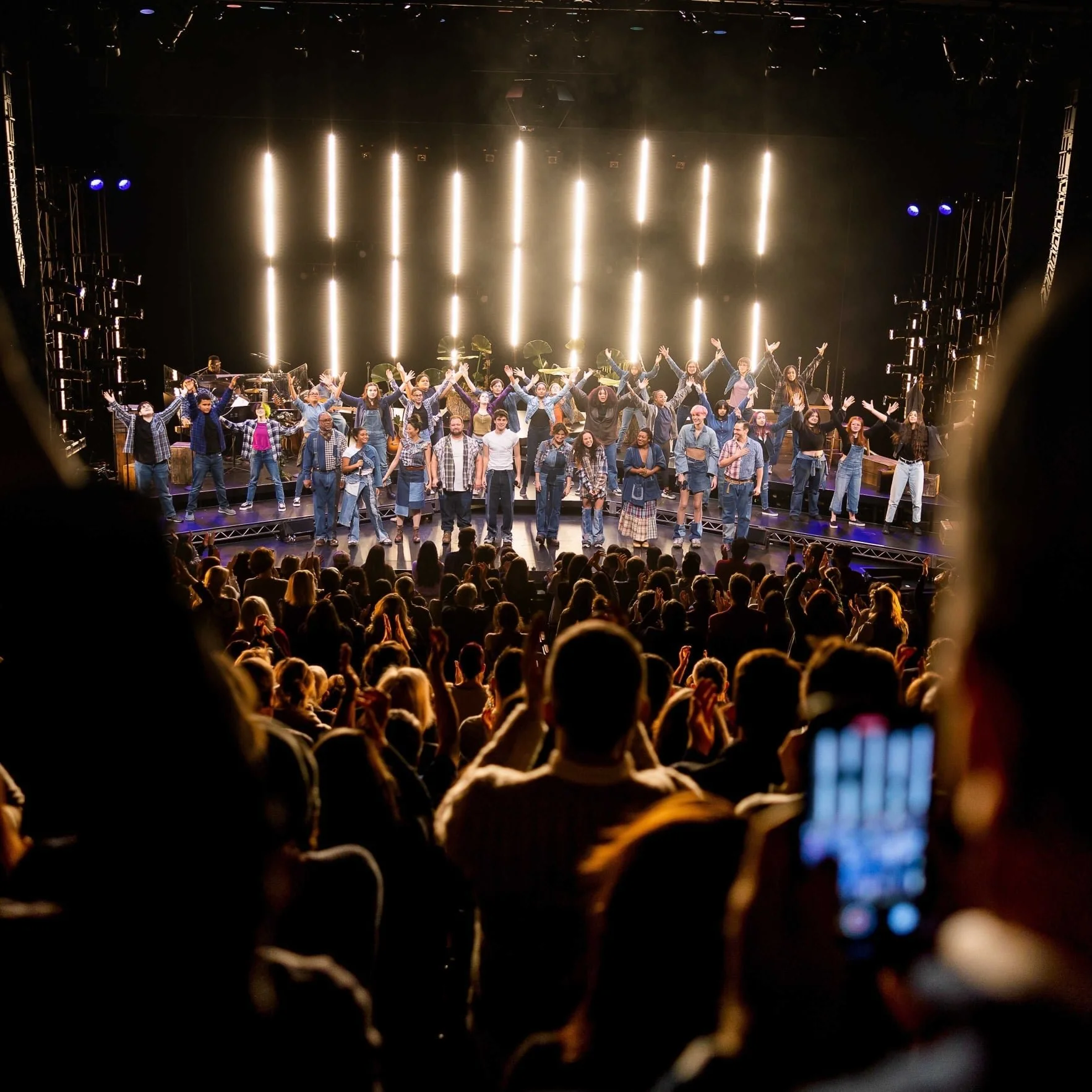 On stage at a concert or theater, a diverse group of performers, including children and adults, are standing with their arms raised in front of illuminated vertical light bars and an audience watching from below.