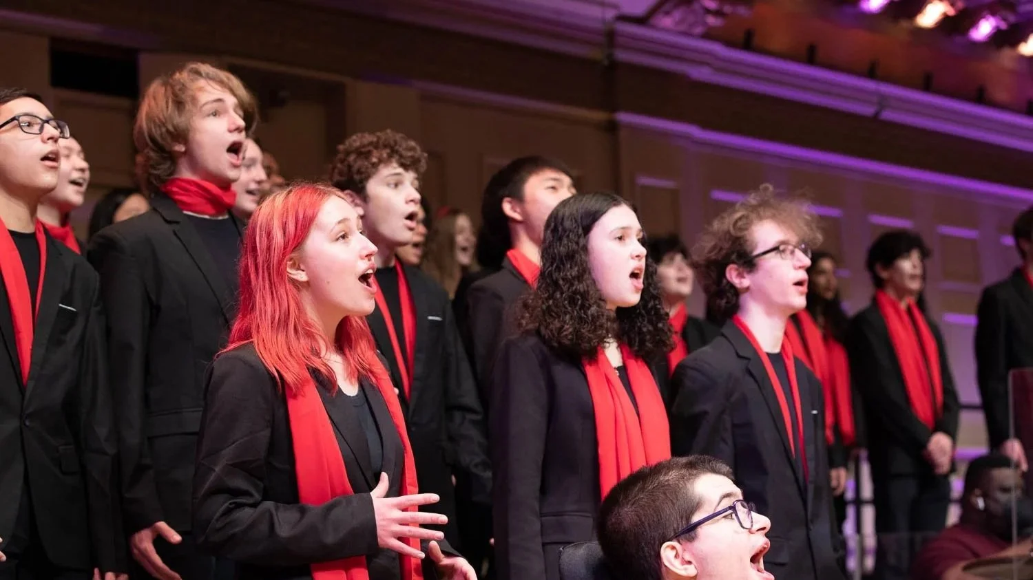 Young boys and girls in formal black suits with red scarves singing in a choir performance.
