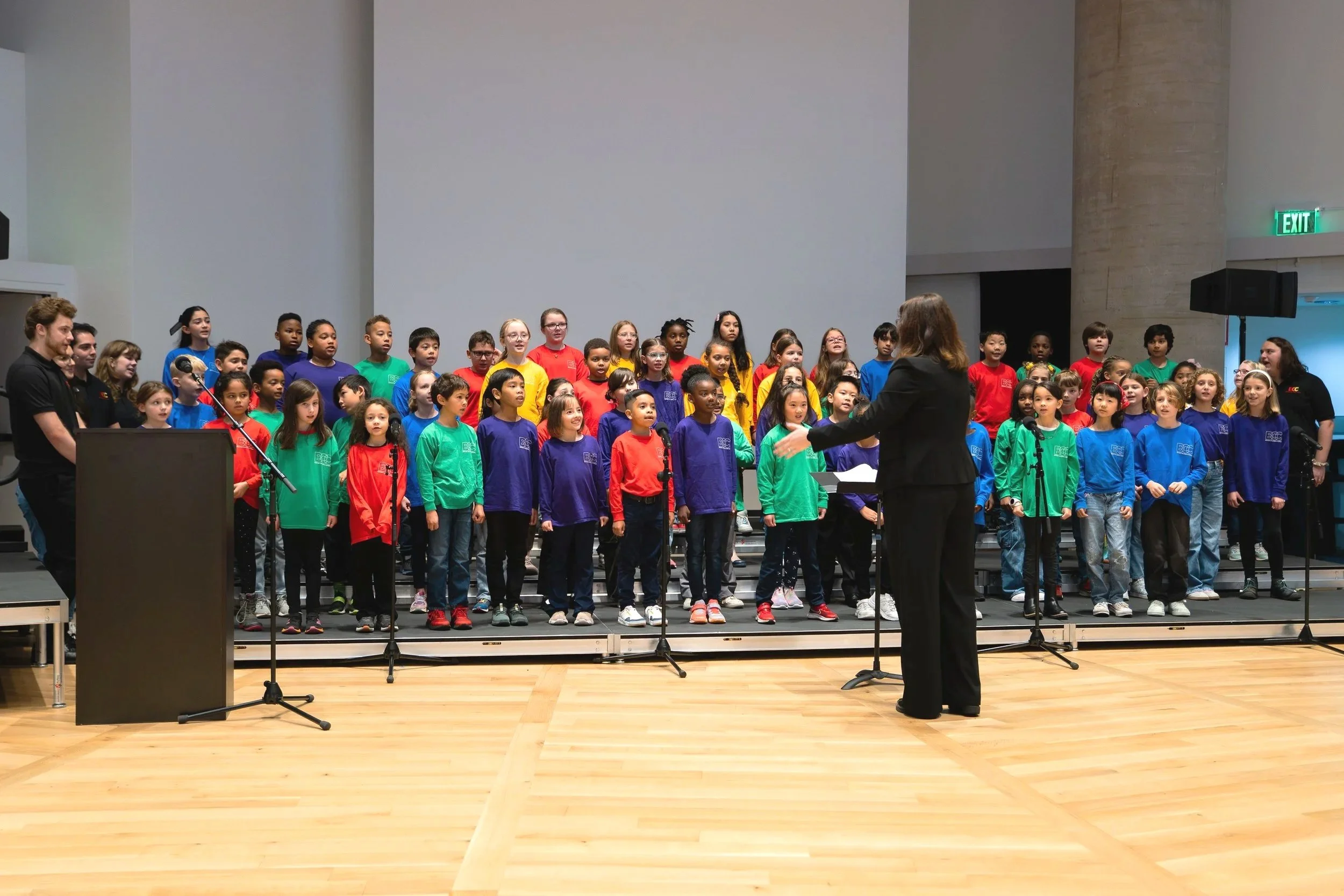 Children in colorful shirts singing on stage, directed by a woman with brown hair, in a concert hall with wooden floors and gray walls.