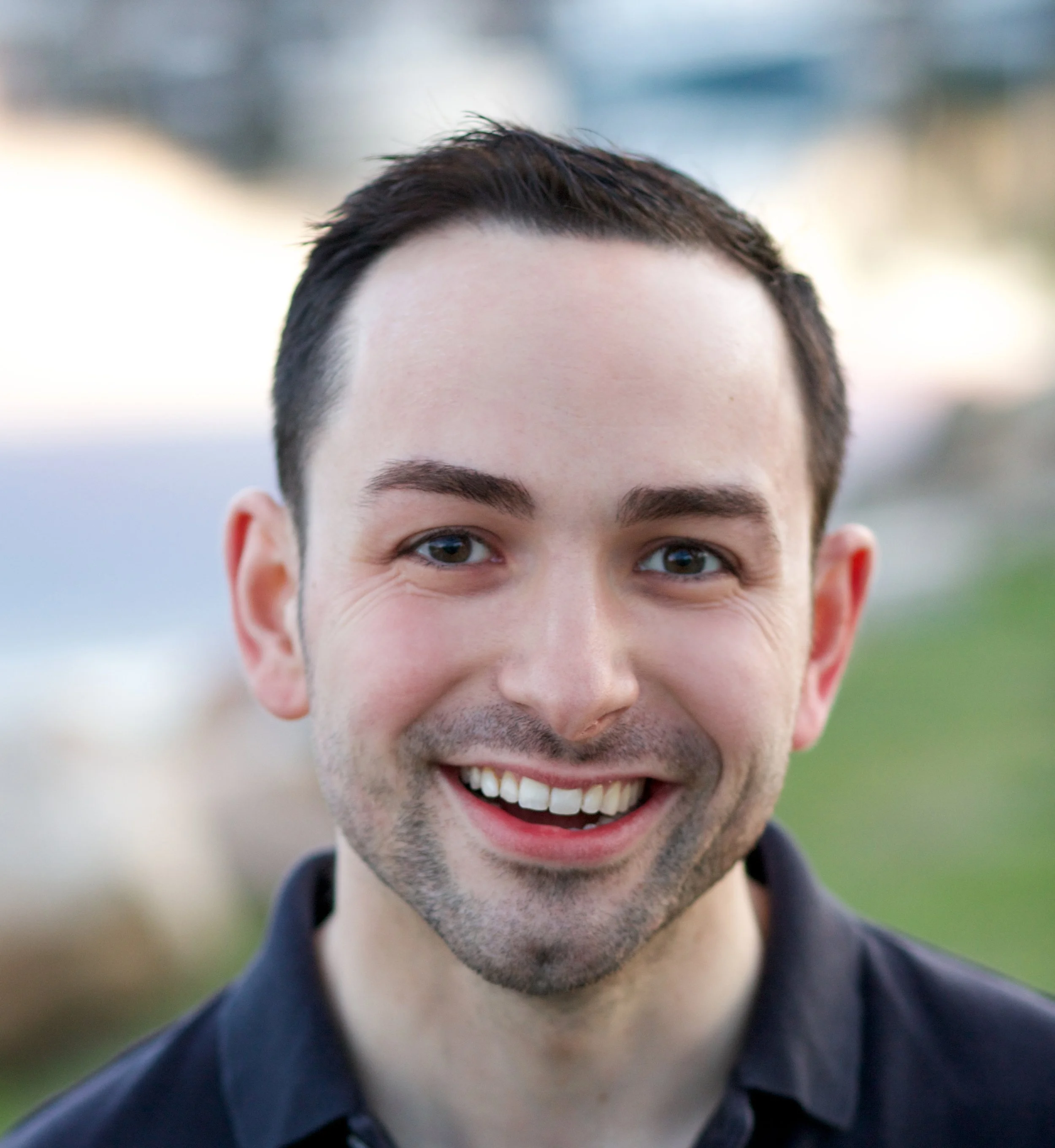 Smiling man with dark hair and blue eyes outdoors, blurred background.