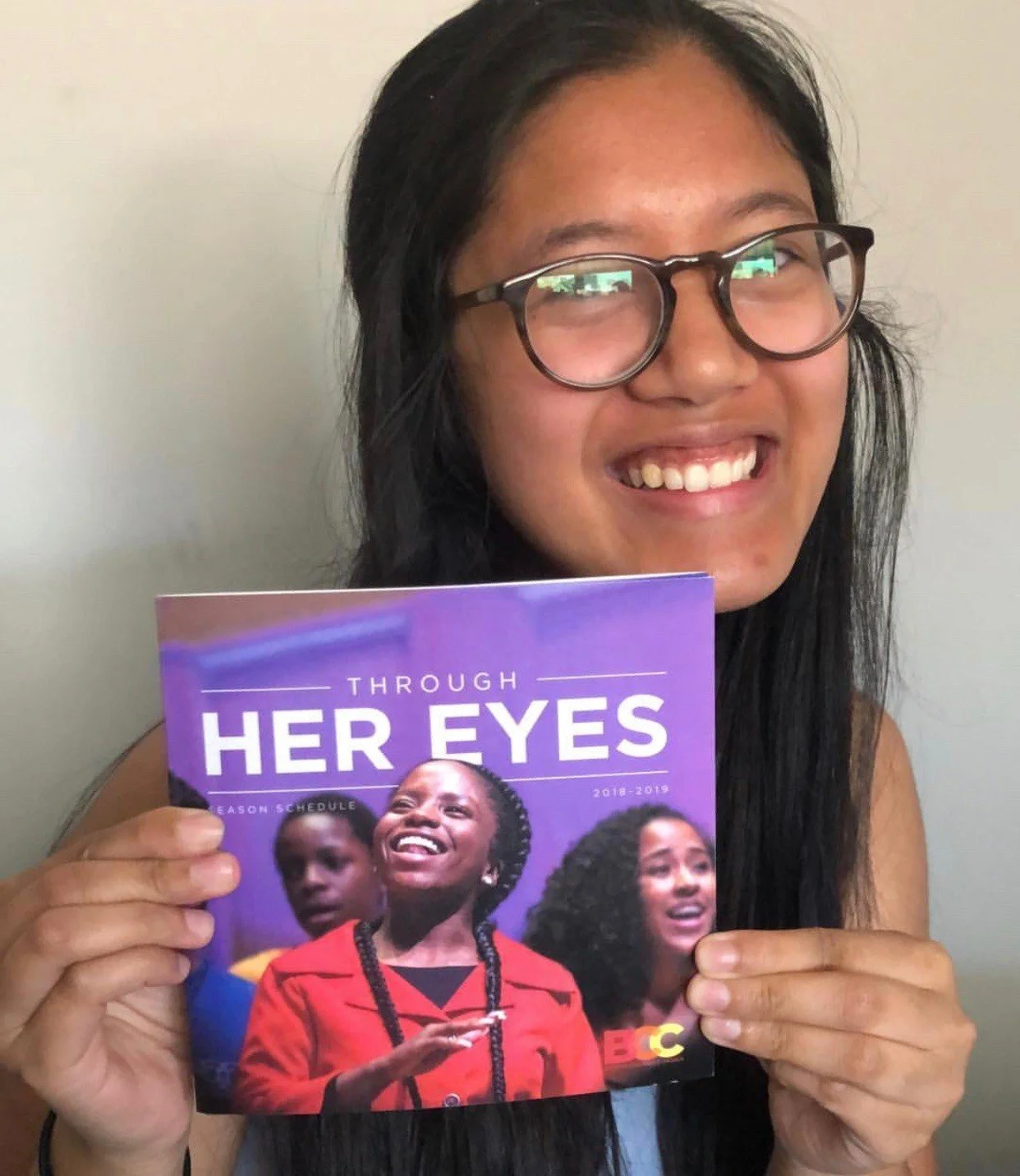 A young woman with glasses smiling and holding a colorful booklet titled 'Through Her Eyes' featuring images of diverse women on the cover.
