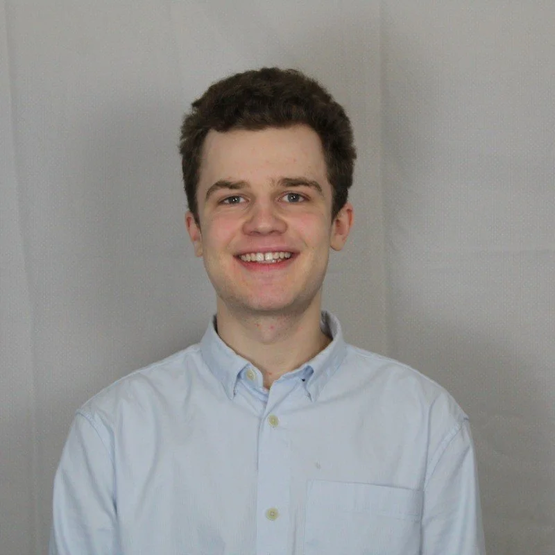A young man with brown hair, wearing a light blue button-up shirt, smiling at the camera, standing against a gray background.