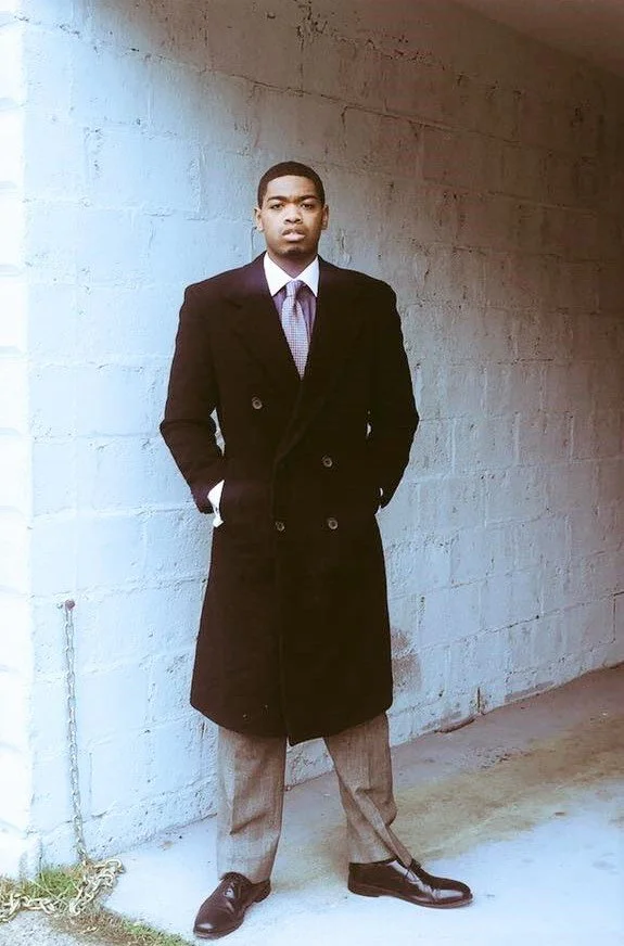 A young man standing outdoors against a white brick wall, wearing a long black coat, a collared shirt, dress pants, and dress shoes.