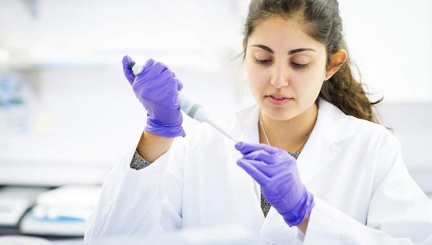 A female scientist wearing a white lab coat and purple gloves carefully using a pipette in a laboratory setting.
