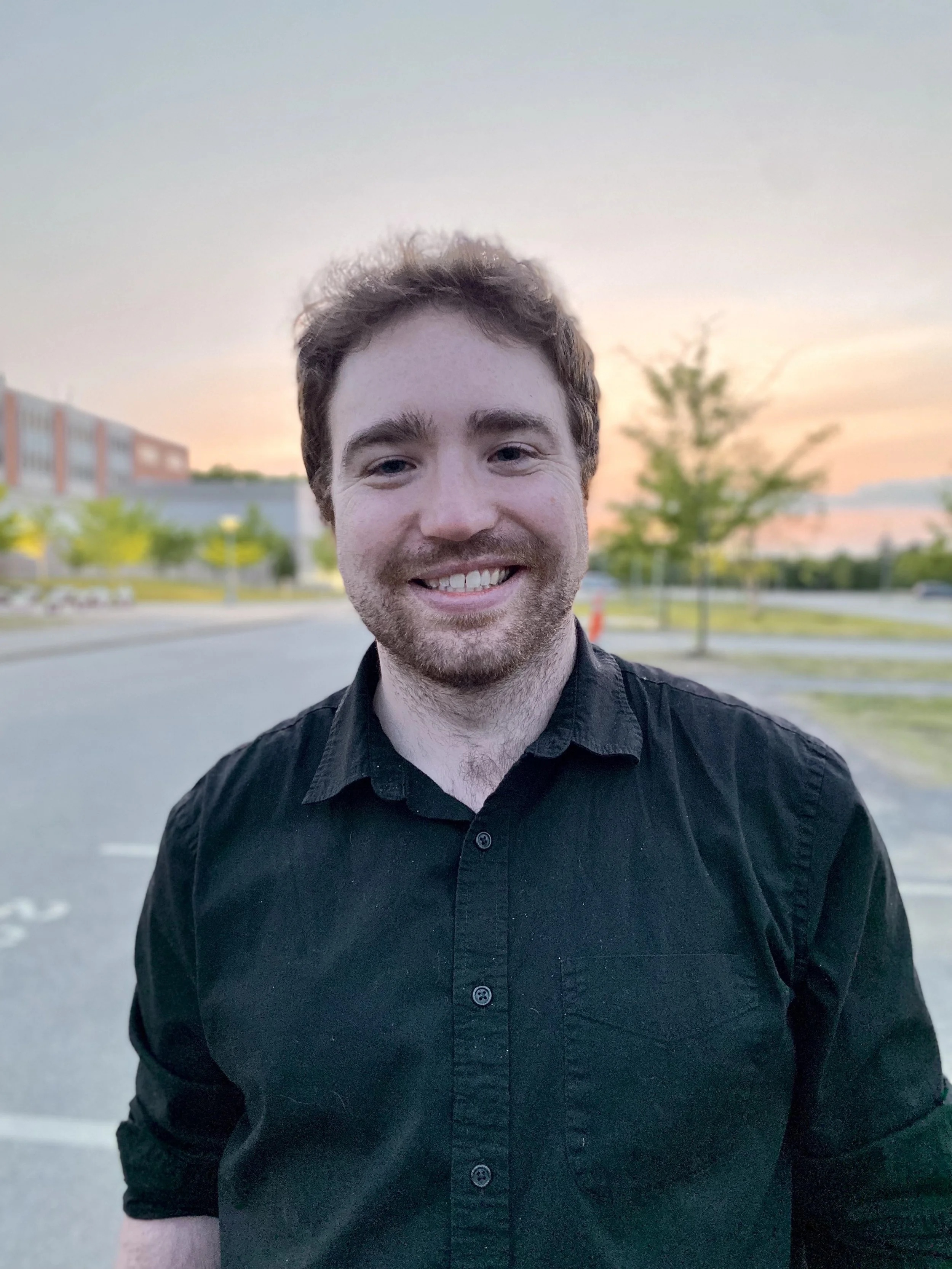 A smiling man wearing a black shirt standing outdoors during sunset with trees, a building, and a parking lot in the background.