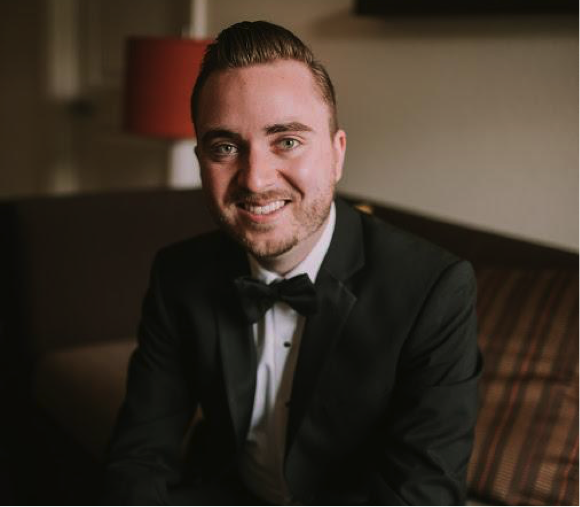 A smiling man in a tuxedo with a bow tie, sitting on a sofa in a warmly lit room.