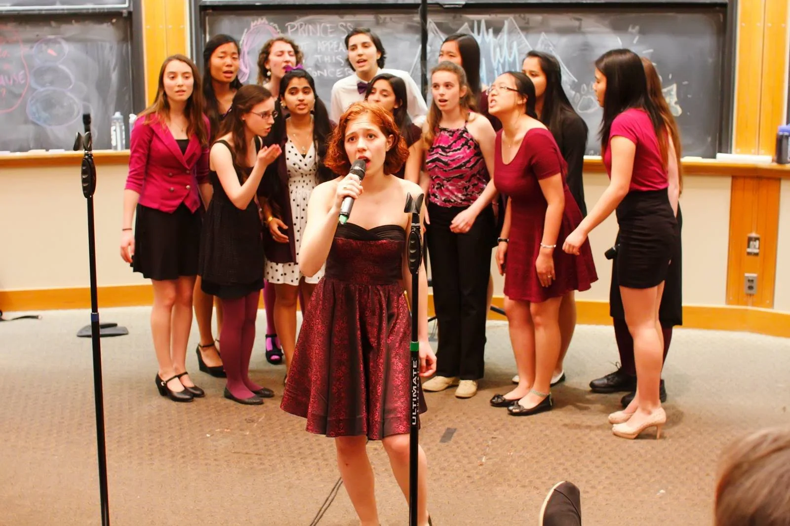A young woman with red hair, wearing a strapless dress, singing into a microphone in front of a group of girls in a classroom setting, with chalkboards in the background.